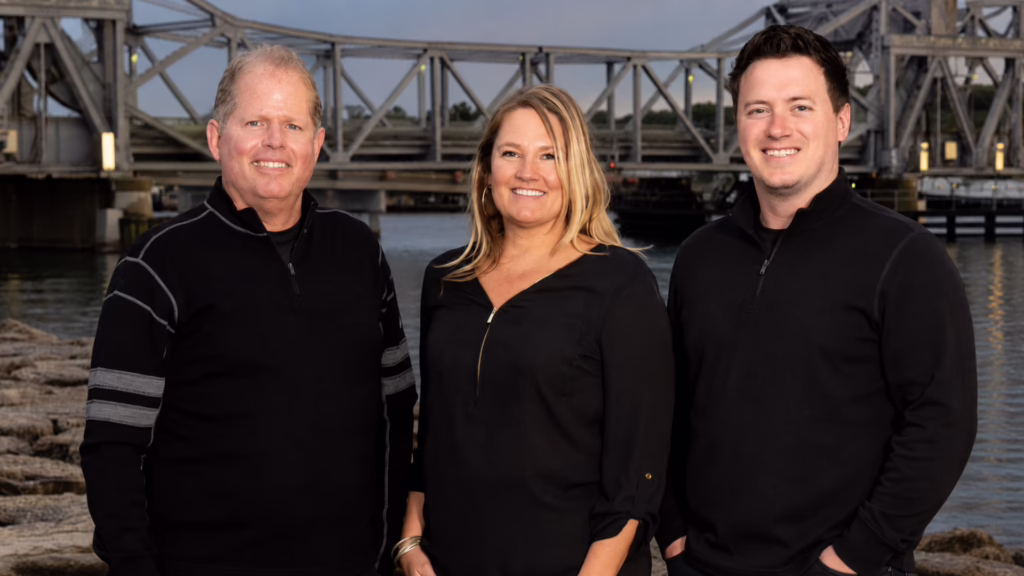 Three adults stand side by side by a riverfront with a metal bridge in the background, smiling at the camera.